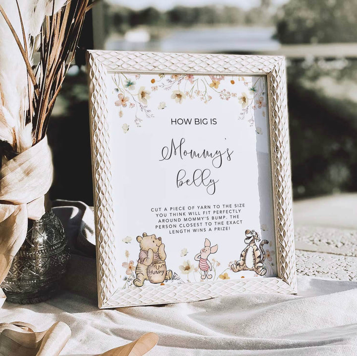 Winnie The Pooh themed "How Big Is Mommy's Belly" game sign on table at baby shower, decorative frame with floral accents.